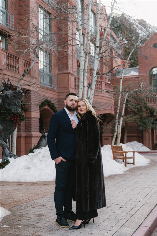 Romantic winter proposal at the St. Regis Aspen featuring a couple kissing under a dramatic red rose floral arch with snow-covered evergreens and Aspen Mountain in the background."