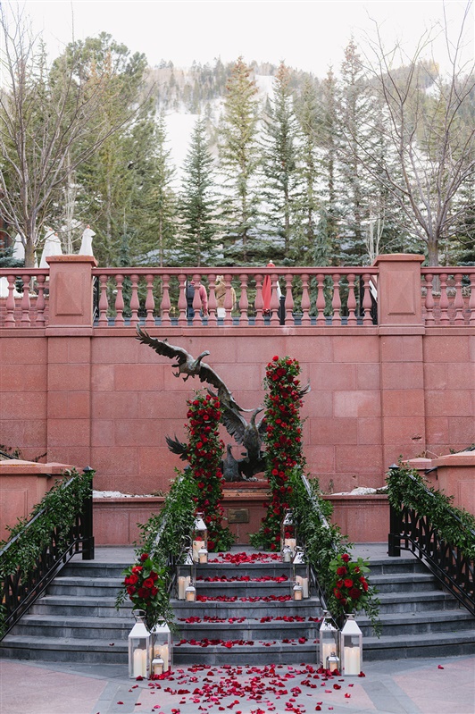 Romantic winter proposal at the St. Regis Aspen featuring a couple kissing under a dramatic red rose floral arch with snow-covered evergreens and Aspen Mountain in the background