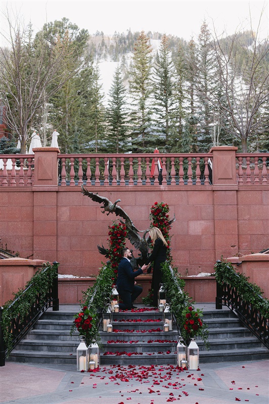 Romantic winter proposal at the St. Regis Aspen featuring a couple kissing under a dramatic red rose floral arch with snow-covered evergreens and Aspen Mountain in the background