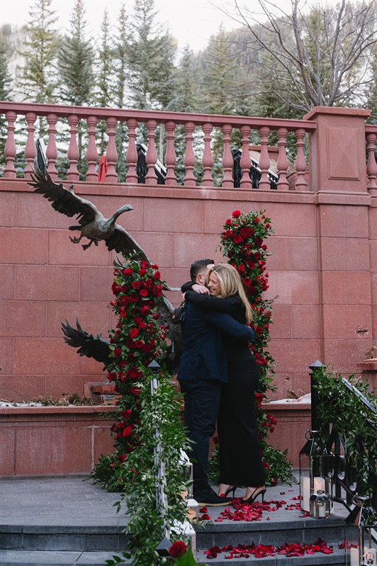 Romantic winter proposal at the St. Regis Aspen featuring a couple kissing under a dramatic red rose floral arch with snow-covered evergreens and Aspen Mountain in the background